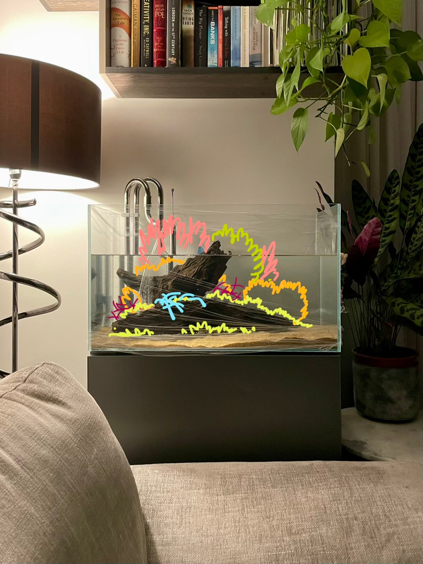 A rectangular glass aquarium with a dark driftwood centerpiece and colorful neon coral-like decorations, displayed on a dark pedestal in a living room setting with a lamp, sofa, and houseplants visible.
